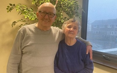 A man with glasses hugging a woman with a large plant behind them