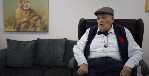 A man wearing a flat cap, a white shirt, a bow tie and a waistcoat with a poppy attached to it sitting in a chair.