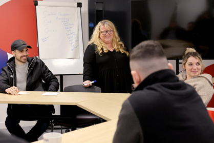 A woman standing and smiling at a desk with three people sitting around her.