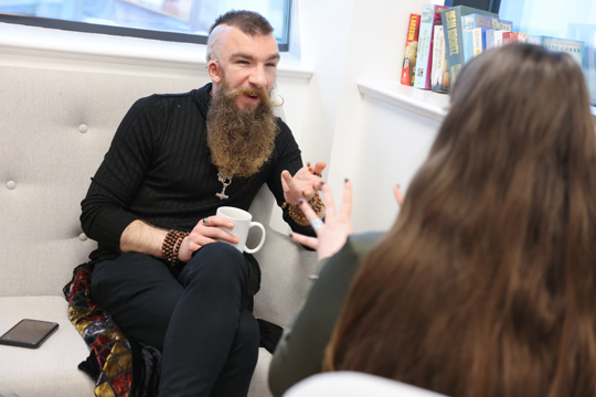 A man with a beard and dressed in black sitting with a cup on his lap and talking to a woman with long dark hiar.