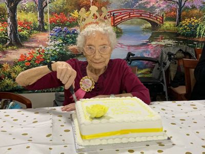 A woman with glasses wearing a crown cutting into her birthday cake
